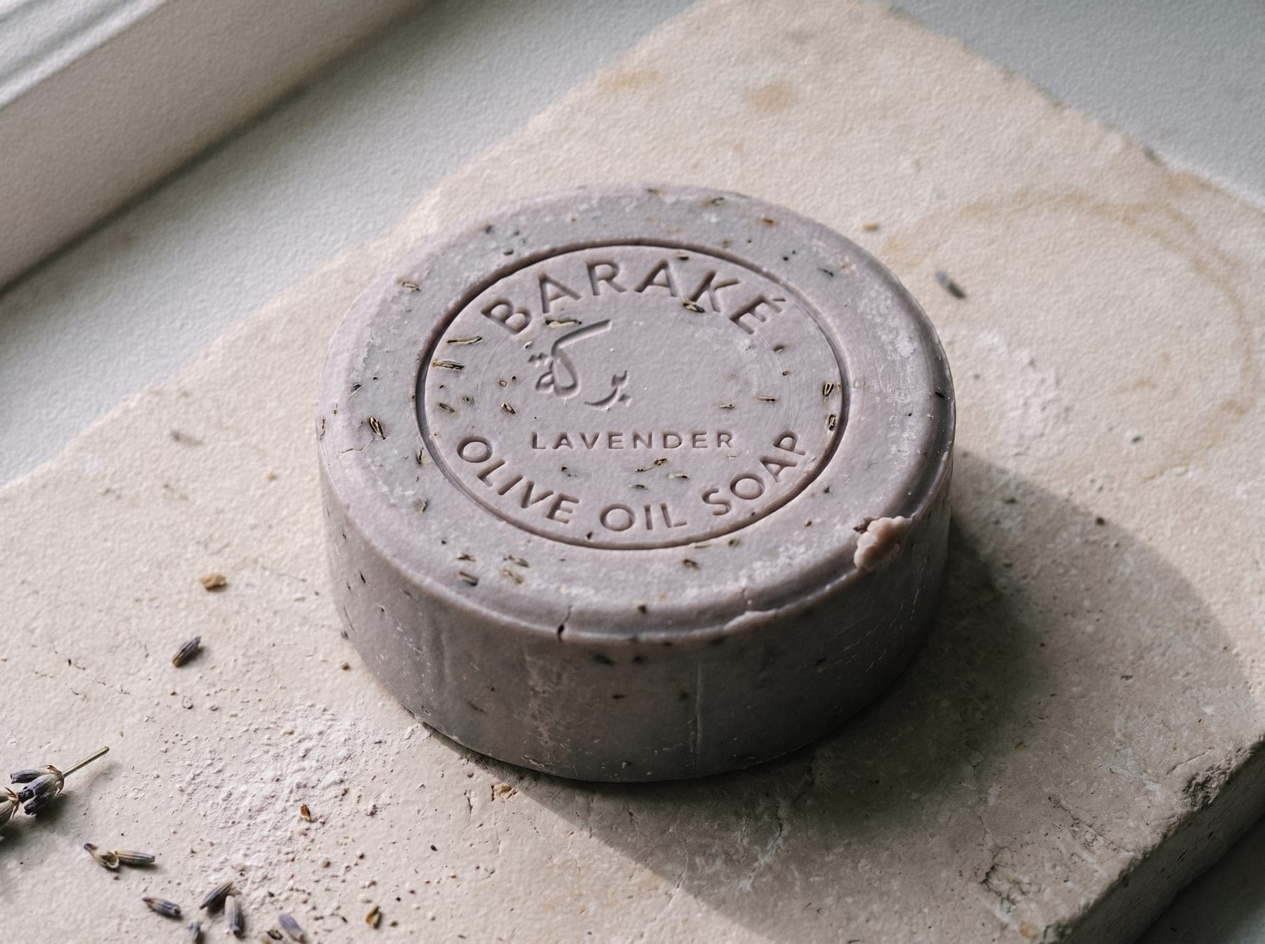 A round Baraké olive oil soap, lavender, flecked with dried buds, with the Baraké seal pressed into the top, on pale stone.
