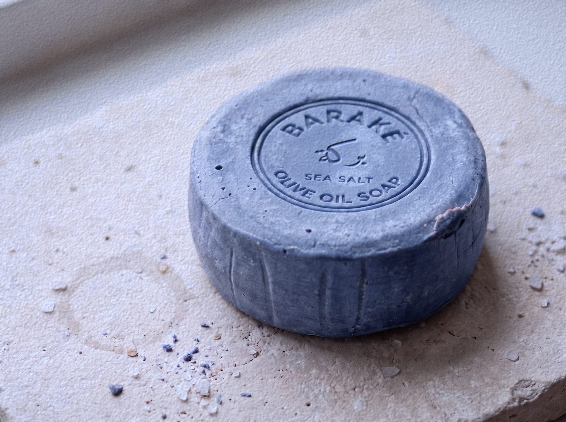 A round Baraké olive oil soap, sea salt, blue grey, with the Baraké seal pressed into the top, on pale stone.